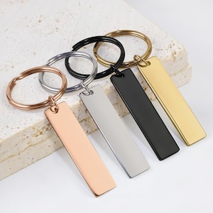 Könnte beinhalten: Eine Sammlung rechteckiger Schlüsselanhänger in verschiedenen Metallausführungen: Roségold, Silber, Schwarz und Gold. Jeder Schlüsselanhänger hat ein schlichtes, elegantes Design mit einem rechteckigen Anhänger, der an einem kreisförmigen Schlüsselring befestigt ist. Die Schlüsselanhänger werden auf einer strukturierten, cremefarbenen Oberfläche präsentiert.