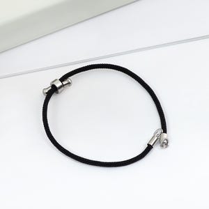 Può includere: Un braccialetto in corda nera con chiusure in metallo color argento. Il braccialetto è regolabile e presenta un design semplice e minimalista. La corda ha un diametro di circa 0,5 cm.