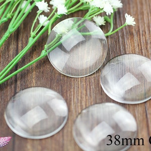 May include: Four clear glass cabochons, each measuring 38mm in diameter, are arranged on a wooden surface. The cabochons are round and have a smooth, polished finish. The text "38mm Glass" is printed in black on the wooden surface.