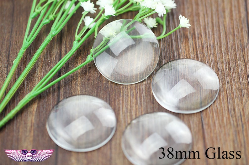May include: Four clear glass cabochons, each measuring 38mm in diameter, are arranged on a wooden surface. The cabochons are round and have a smooth, polished finish. The text "38mm Glass" is printed in black on the wooden surface.