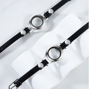 Könnte beinhalten: Zwei schwarze Lederarmbänder mit silbernen runden Anhänger. Die Anhänger haben klare Glaseinsätze.