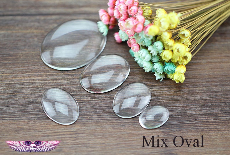 Pode incluir: Uma cole&ccedil;&atilde;o de caboch&otilde;es de vidro transparentes em forma oval e em v&aacute;rios tamanhos. Os caboch&otilde;es est&atilde;o dispostos sobre uma superf&iacute;cie de madeira, com um pequeno buqu&ecirc; de flores secas coloridas ao fundo. O texto "Mix Oval" &eacute; vis&iacute;vel.