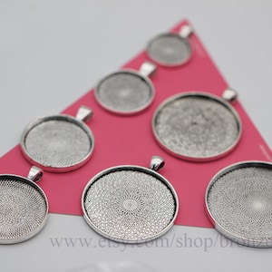 May include: A set of eight silver-toned round pendant trays with a decorative patterned design. The trays are arranged on a pink surface.