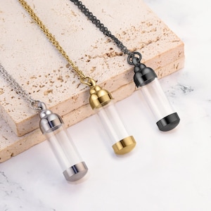 Puede incluir: Tres collares con viales de vidrio y cadenas de metal. Los viales son transparentes, y las tapas y cadenas son plateadas, doradas y negras. Los collares se exhiben sobre una superficie clara y texturizada.