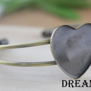 Puede incluir: Una pulsera de metal dorado con una banda doble y un dije de vidrio en forma de corazón. El dije tiene una superficie lisa y negra y un borde festoneado.