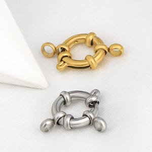 Puede incluir: Dos cierres metálicos, uno dorado y otro plateado, se muestran sobre un fondo blanco. Cada cierre presenta un diseño circular con detalles anudados redondeados y pequeños bucles para la fijación. Estos cierres probablemente se utilizan para la fabricación de joyas.