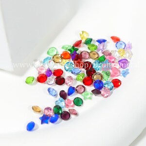 May include: Assortment of colorful crystal rhinestones in various shapes, including stars, hearts, and rounds. The rhinestones are scattered on a white surface.