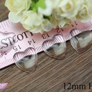 May include: Four clear heart-shaped glass beads, each 12mm in size, are displayed on a wooden surface. One heart is pink with the word "WESTCOTT" printed on it. A pink ruler and white flowers are in the background.