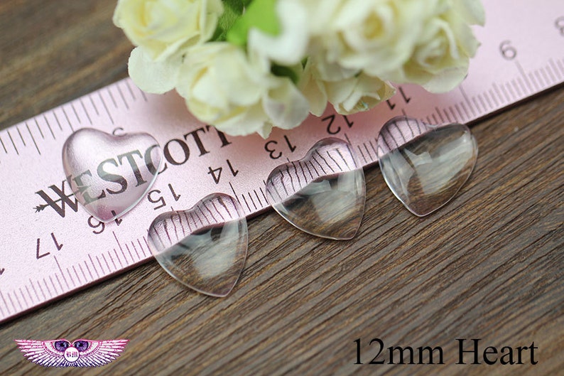 May include: Four clear heart-shaped glass beads, each 12mm in size, are displayed on a wooden surface. One heart is pink with the word "WESTCOTT" printed on it. A pink ruler and white flowers are in the background.