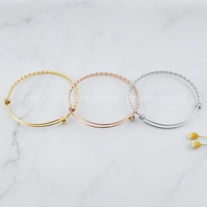 May include: Three braided bangle bracelets in gold, rose gold, and silver. The bracelets are adjustable and have a simple, elegant design.