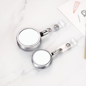 May include: Two silver retractable badge holders with clear plastic straps. Each holder has a round, blank white face and a metal clip. The badge holders are on a white marble surface.
