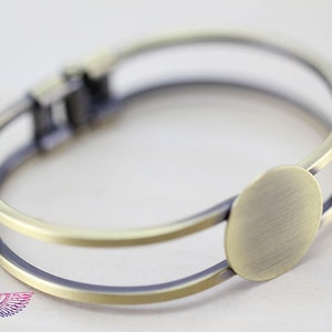 May include: A gold-toned metal bangle bracelet with a round, flat, blank space for customization. The bracelet is double-layered and has a hinged clasp.