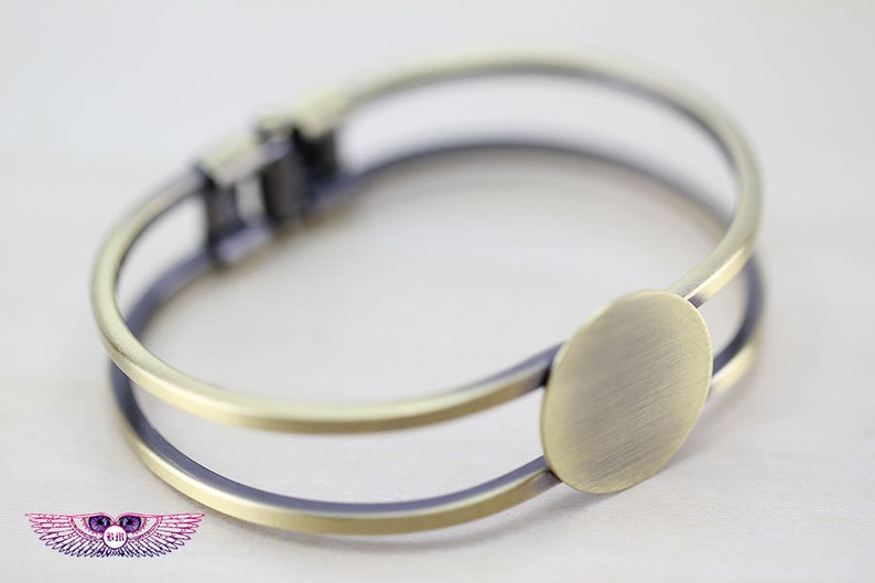 May include: A gold-toned metal bangle bracelet with a round, flat, blank space for customization. The bracelet is double-layered and has a hinged clasp.