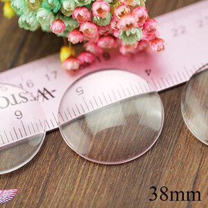 May include: Two clear glass circles, each measuring 38 mm in diameter, are placed on a wooden surface. A pink ruler with black markings is partially visible behind the circles.