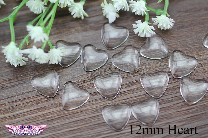 May include: Clear, heart-shaped glass beads, each measuring 0.47 inches, are scattered on a wooden surface. White flowers and greenery are in the background. The image includes the text "12mm Heart".