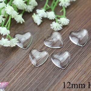 May include: Five clear glass heart-shaped embellishments, each measuring 12mm, are arranged on a wooden surface. Delicate white flowers and green stems are in the background. The text "12mm Heart" is visible.