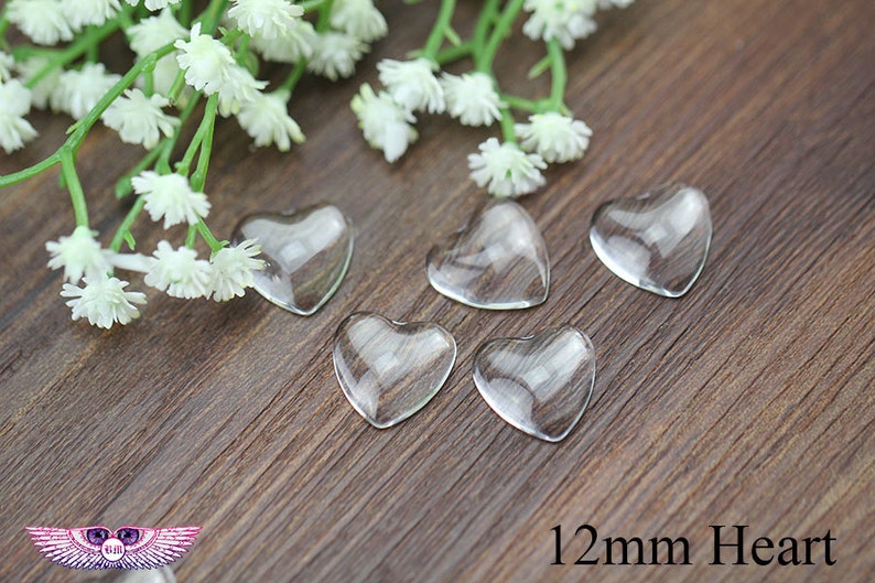 May include: Five clear glass heart-shaped embellishments, each measuring 12mm, are arranged on a wooden surface. Delicate white flowers and green stems are in the background. The text "12mm Heart" is visible.