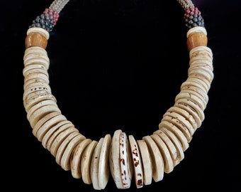 May include: Cream-colored disc necklace with a woven beige cord. The necklace features large, flat, circular beads with brown markings. The cord transitions to smaller black and red beads near the clasp, with wooden beads as accents. The necklace is displayed against a black background.