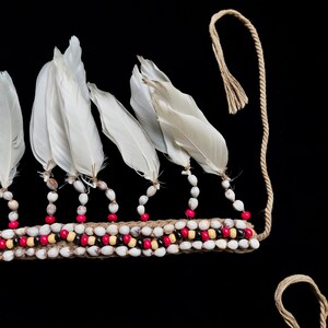 May include: A detailed view of a headpiece with white feathers and a beaded band. The band is adorned with white, red, and black beads, and is connected to a natural fibre cord. The feathers are attached to the band with small beads.