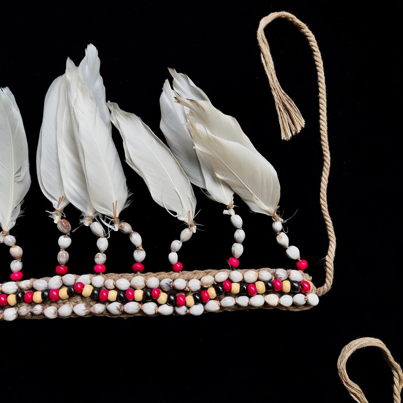 May include: A detailed view of a headpiece with white feathers and a beaded band. The band is adorned with white, red, and black beads, and is connected to a natural fibre cord. The feathers are attached to the band with small beads.
