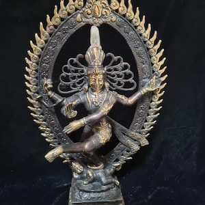 Könnte beinhalten: Bronzestatue einer Gottheit in dynamischer Pose, umrahmt von einem verzierten, goldenen Heiligenschein. Die Figur ist mit detailliertem Schmuck und einer hohen Kopfbedeckung geschmückt. Die Statue steht auf einem quadratischen Sockel, mit dunklem Hintergrund.