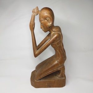 Peut inclure: Sculpture en bois représentant une personne agenouillée, les mains jointes en prière. La figure a une expression sereine et est ornée d'une coiffe florale. La sculpture est sculptée dans un bois brun clair et repose sur une base rectangulaire.