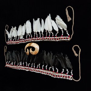 May include: Two decorative headbands with white and black feathers, decorated with red, white, and tan beads. A tan, spiral-shaped pendant is centered between the headbands. The background is solid black.