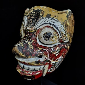 Könnte beinhalten: Eine Holzmaske mit einem grimmigen Ausdruck, bemalt in Rot, Schwarz, Weiß und Gelb. Die Maske hat große Augen, einen breiten Mund mit Zähnen und komplizierte Details.