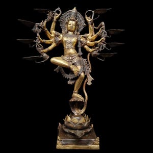 May include: A golden statue of a deity with multiple arms, adorned with intricate details. The figure is posed gracefully, standing on a lotus flower base. The statue has a halo and holds objects, with lamps extending from the back.