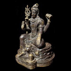 Puede incluir: Una estatua de bronce de la deidad hindú Shiva, sentado con las piernas cruzadas y sosteniendo un tridente en una mano y un tambor en la otra. Está adornado con joyas y una guirnalda de flores. La estatua está colocada sobre una base con una representación de un león.