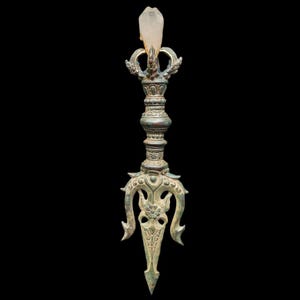 May include: An antique bronze-colored ritual object with intricate carvings and a clear crystal at the top. The object has a trident-like base and a detailed central shaft. The background is black.