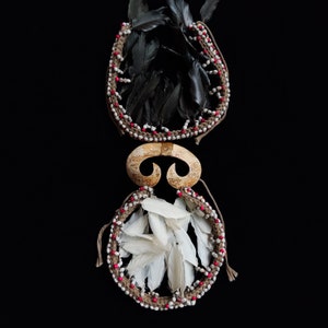 May include: Two handcrafted tribal ornaments. The top ornament features black feathers within a woven frame adorned with white and red beads. The bottom ornament has white feathers and a similar frame. A curved, bone-like piece connects the two.