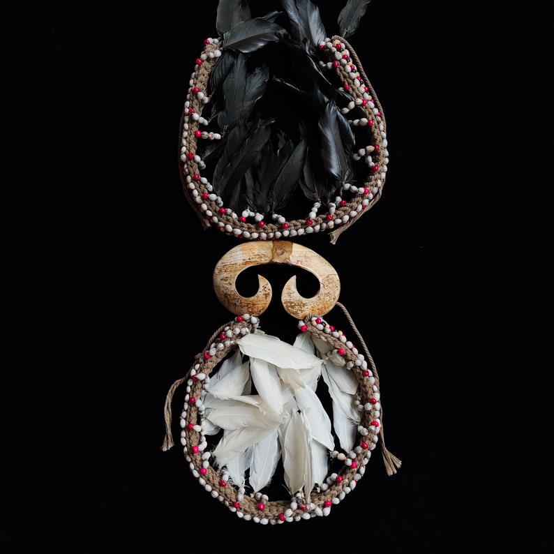 May include: Two handcrafted tribal ornaments. The top ornament features black feathers within a woven frame adorned with white and red beads. The bottom ornament has white feathers and a similar frame. A curved, bone-like piece connects the two.