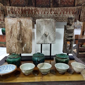 May include: A display of antique Asian artefacts, including carved wooden panels, a wooden statue, ceramic bowls, and lidded jars in shades of green and brown. The items are arranged on a glass-topped table, with a decorative wooden structure in the background.
