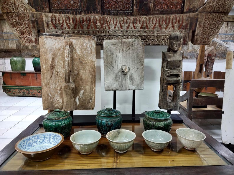 May include: A display of antique Asian artefacts, including carved wooden panels, a wooden statue, ceramic bowls, and lidded jars in shades of green and brown. The items are arranged on a glass-topped table, with a decorative wooden structure in the background.