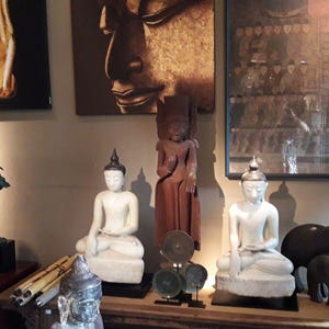 May include: A collection of Asian-inspired sculptures and artwork. The display includes white and brown Buddha statues, a bronze-colored statue, and paintings of hands and a Buddha face. The items are arranged on a wooden surface.