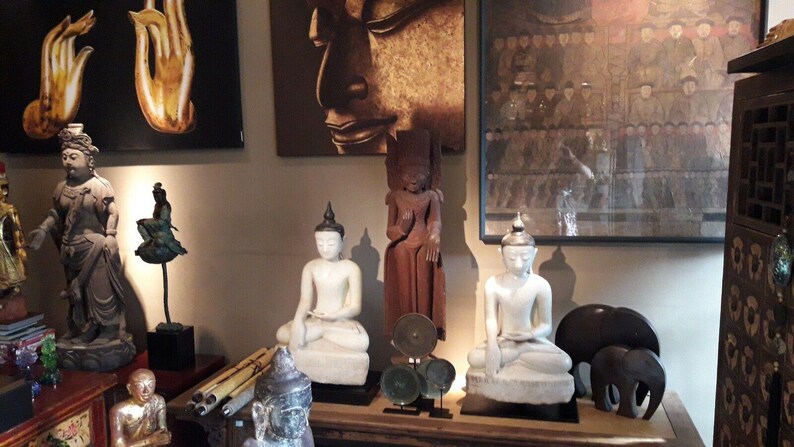 May include: A collection of Asian-inspired sculptures and artwork. The display includes white and brown Buddha statues, a bronze-colored statue, and paintings of hands and a Buddha face. The items are arranged on a wooden surface.