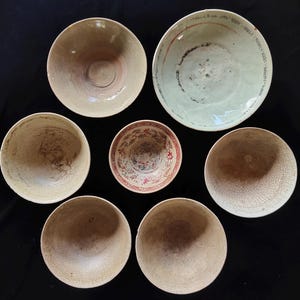 以下が含まれることがあります： 7つの陶器のボウルがグループで配置されています。セットには、無地のベージュやひび割れた質感のものから、中央にある赤と青の複雑な装飾模様やモチーフが施されたボウルまで、さまざまな釉薬仕上げが含まれています。