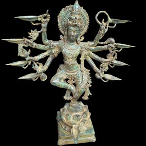 May include: A bronze-toned metal statue of a multi-armed deity standing on a lotus base. The figure has ten arms, each holding a small oil lamp, and wears an ornate crown and jewelry. The surface has a weathered, aged patina finish.