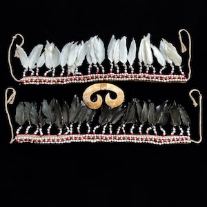 May include: Two feather necklaces with beaded detailing and a bone pendant. One necklace has white feathers, the other black. The beads are white and red. The pendant is a light tan colour and has a curved shape.