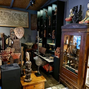 May include: A shop interior showcasing a collection of handcrafted wooden sculptures and carvings. The display includes statues, masks, and decorative objects of various sizes and styles. A large framed artwork is mounted on the wall.