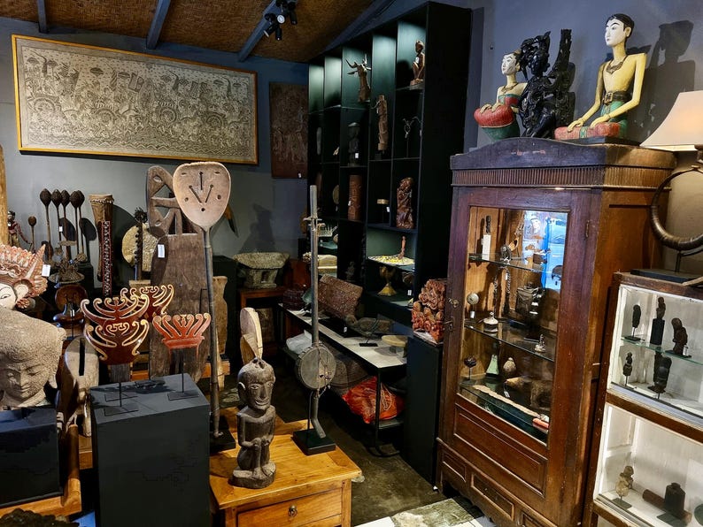 May include: A shop interior showcasing a collection of handcrafted wooden sculptures and carvings. The display includes statues, masks, and decorative objects of various sizes and styles. A large framed artwork is mounted on the wall.