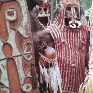 May include: A person in traditional tribal dress, with a woven red and white vest and head covering. Beside him, a man holds a decorated shield. The outfit includes a gourd with feathers and beads, representing cultural heritage.