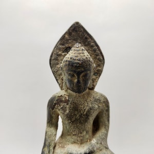 Puede incluir: Una estatua de bronce desgastada de un Buda sentado con una cabeza puntiaguda y una expresión serena. La estatua tiene un halo decorativo detrás de la cabeza.