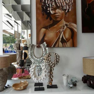 May include: Shell necklaces on black stands are displayed below a portrait of a person with an unusual hairstyle. A geometric mirror is on the left. The necklaces are a mix of white and brown shells.