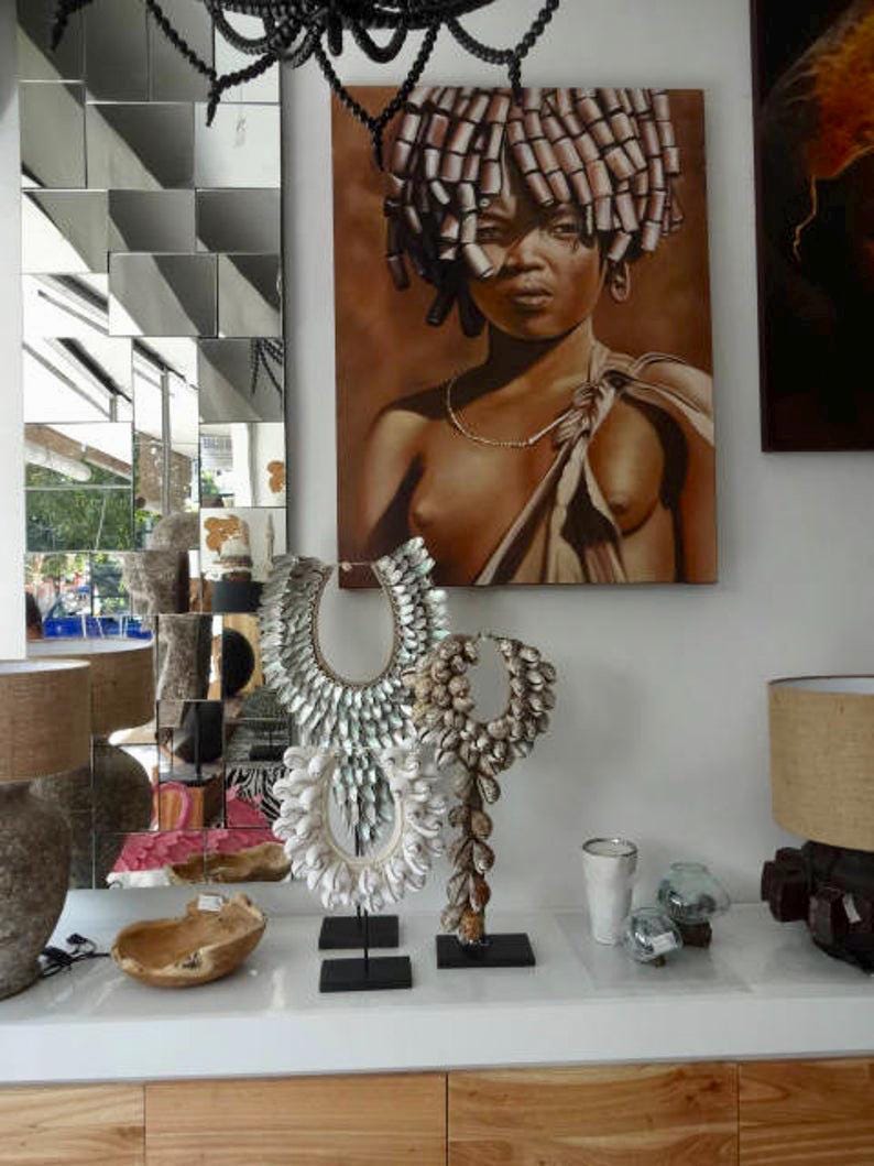 May include: Shell necklaces on black stands are displayed below a portrait of a person with an unusual hairstyle. A geometric mirror is on the left. The necklaces are a mix of white and brown shells.