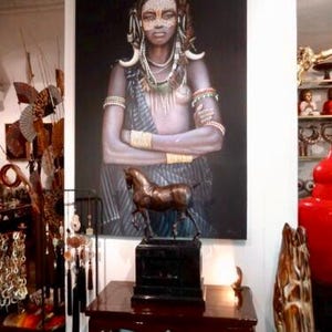 May include: A portrait of a woman with tribal markings and jewellery, displayed above a bronze horse sculpture. The sculpture is on a black base, placed on a dark wood table. Other decorative items are visible in the background.