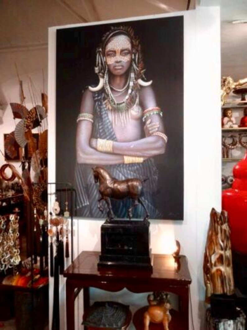 May include: A portrait of a woman with tribal markings and jewellery, displayed above a bronze horse sculpture. The sculpture is on a black base, placed on a dark wood table. Other decorative items are visible in the background.