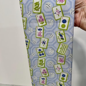 May include: A light blue fabric with a repeating pattern of Mahjong tiles, dice, and a geometric design. The tiles feature various symbols, numbers, and the word "JOKER". The fabric is held up, showcasing the design.