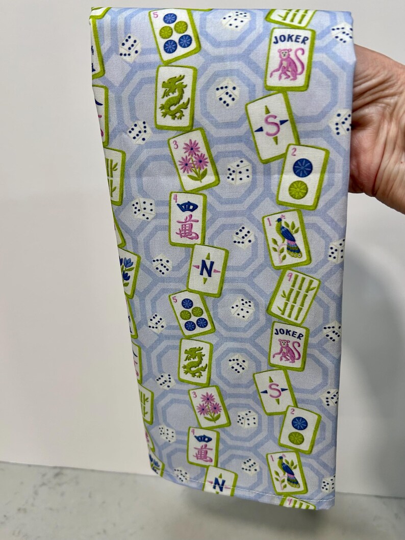 May include: A light blue fabric with a repeating pattern of Mahjong tiles, dice, and a geometric design. The tiles feature various symbols, numbers, and the word "JOKER". The fabric is held up, showcasing the design.